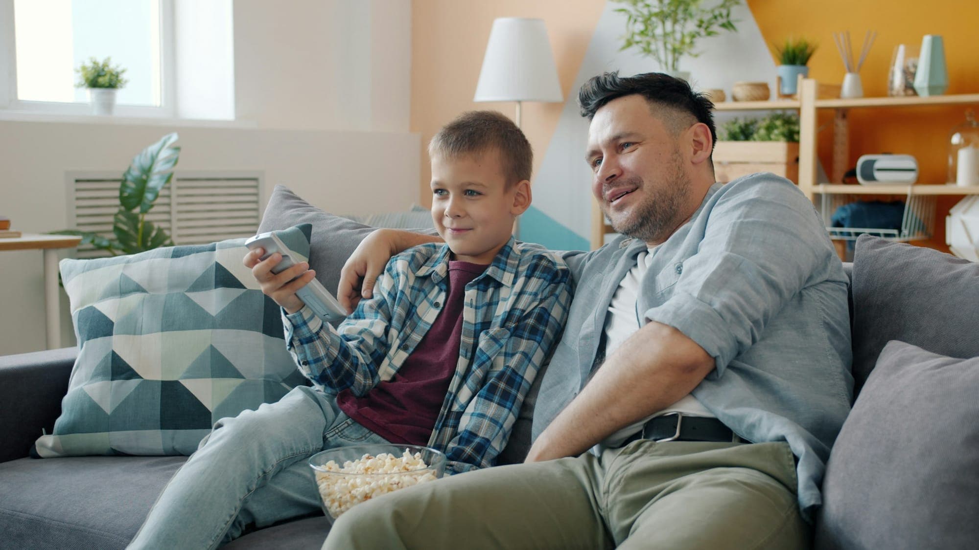 A father and young son on a couch watching television together with a bowl of popcorn