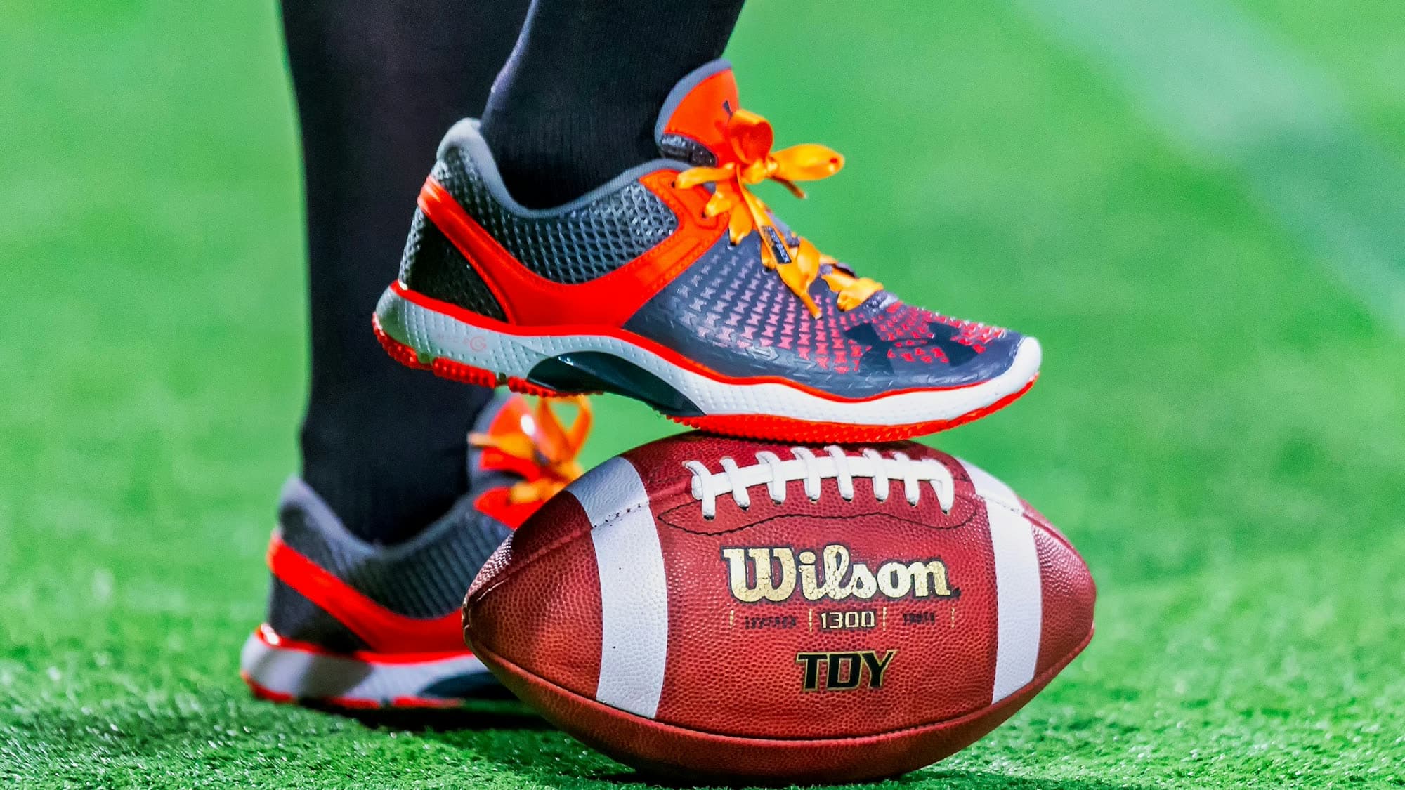 Close-up of a player's cleats resting on a Wilson football on a green turf field