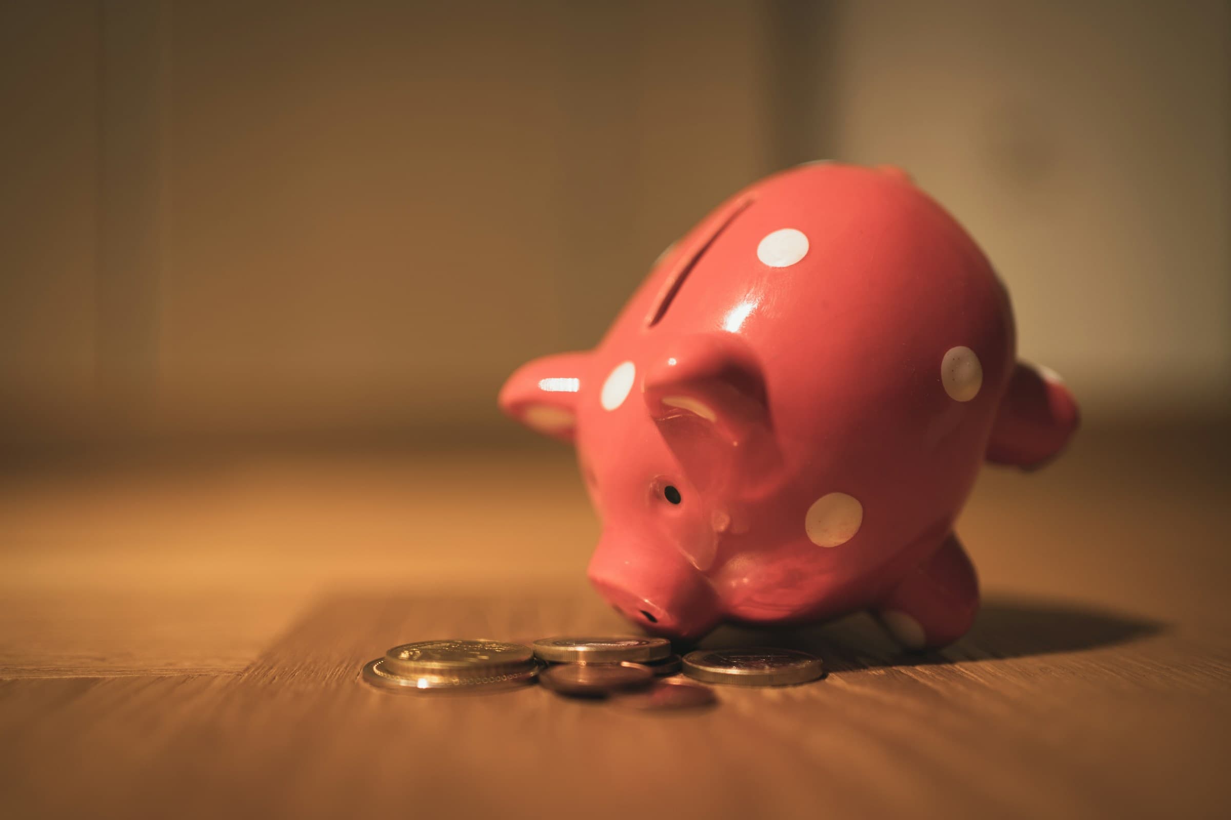 A pink piggy bank next to a small pile of coins on a wooden surface