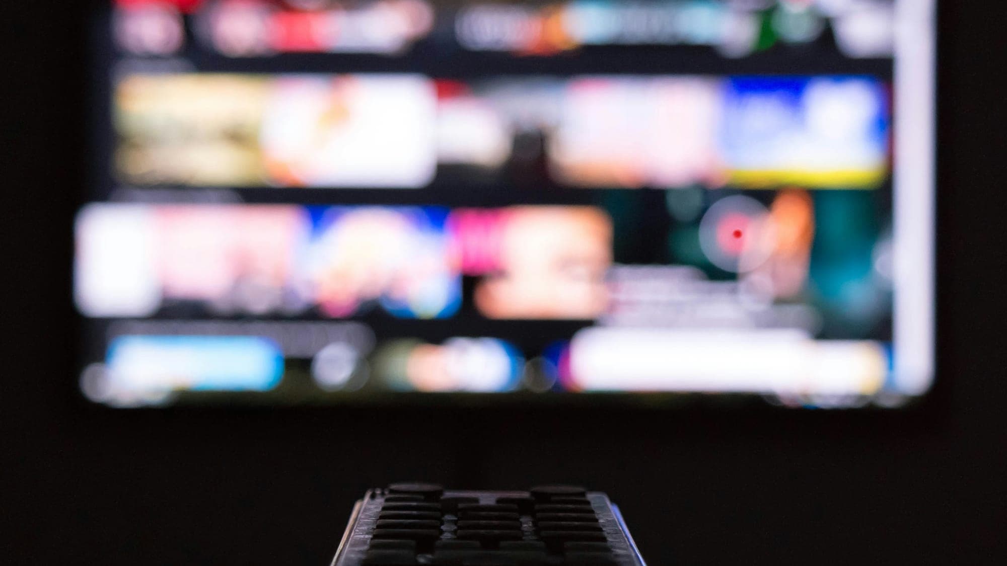 A hand holding a remote control in front of a blurred streaming service menu on a TV