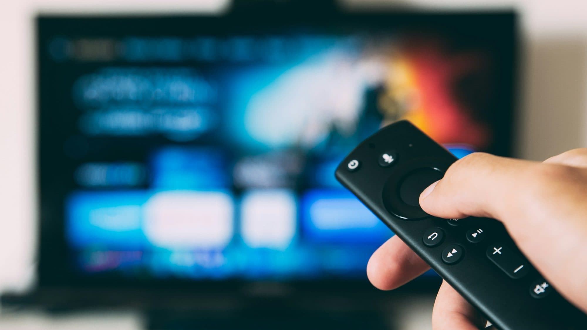 A hand pointing a streaming remote at a TV showing a streaming service's home screen