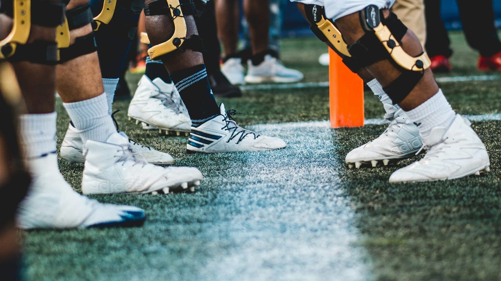 Close-up of college football players' knee braces and cleats lined up at the line of scrimmage next to an orange down marker